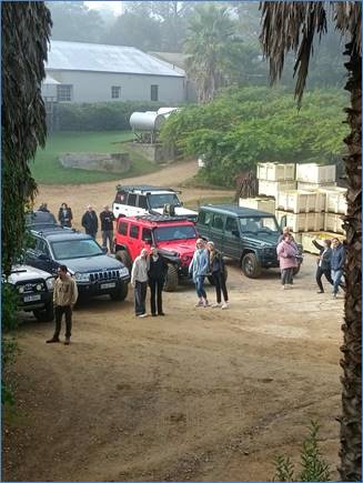 A group of people standing in a dirt area with cars

Description automatically generated